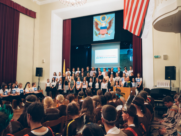 Eine Gruppe von Menschen steht vor einem sitzenden Publikum an einem Podium mit Mikrofon, flankiert von Fahnen und Lautsprechern, während einer zweiten Woche eines Kunst- und Kulturereignisses.