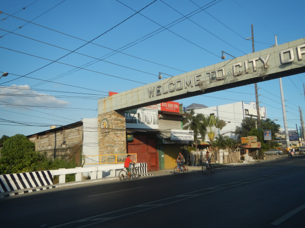 Eine Gruppe von Menschen, die auf Fahrrädern eine Straße in Manila entlangfahren, mit Gebäuden, Bäumen und Strommasten an den Seiten, unter einem bewölkten Himmel und einem "Willkommen in der Stadt Manila"-Bogen.
