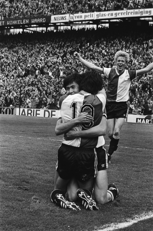 Drei Fussballspieler umarmen sich auf dem Boden, feiernd nach einem Tor vor einer jubelnden Menge in einer schwarz-weissen Stadion-Szene.