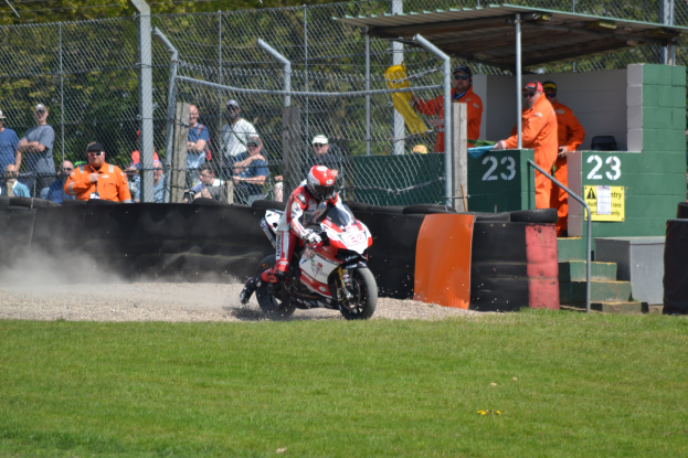 Eine Person in Helm und Handschuhen fährt ein Motorrad über ein grasbewachsenes Feld, mit Zuschauern in Kappen und Brillen, Maschendrahtzaun, Treppe mit Geländer und einem nummerierten Schuppen im Hintergrund.
