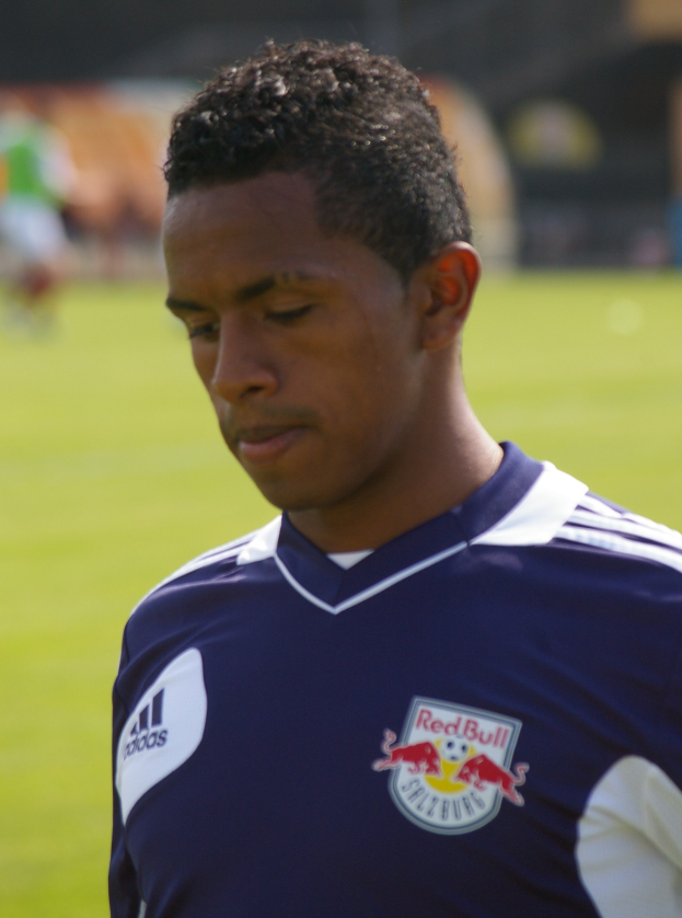 Ein junger Mann in einem blauen T-Shirt auf einem Fußballfeld, der wie ein Flügelstürmer mit kurzen dunklen Haaren und entschlossener Miene aussieht.