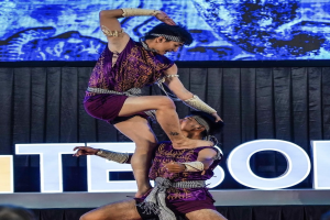 Zwei Akrobaten führen auf der Bühne eine Darbietung auf, wobei der Mann die Frau in seinen Armen hält, während beide purpurfarbene Kleider tragen.