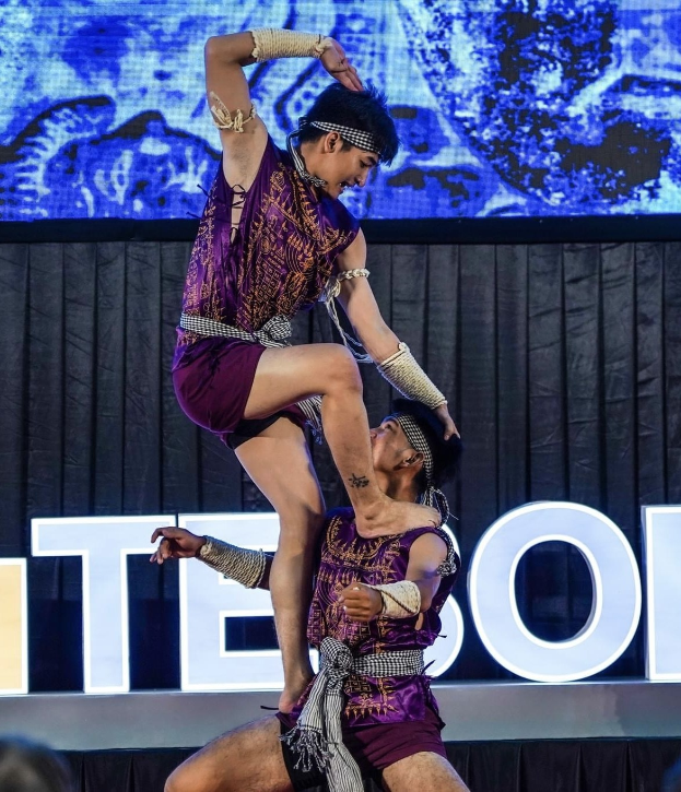 Zwei Akrobaten führen auf der Bühne eine Darbietung auf, wobei der Mann die Frau in seinen Armen hält, während beide purpurfarbene Kleider tragen.