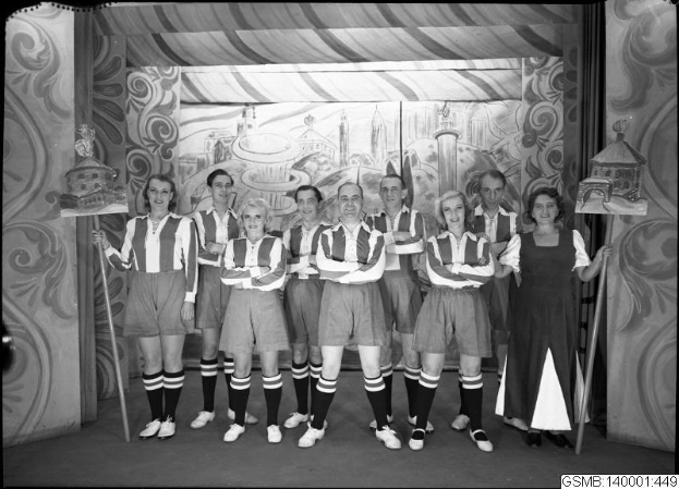 Schwarzes und weißes Foto einer Frauenfußballmannschaft der 1940er Jahre, die auf einer Bühne stehen, lächeln und Stöcke halten, mit einer gemalten Kulisse und Text unten.