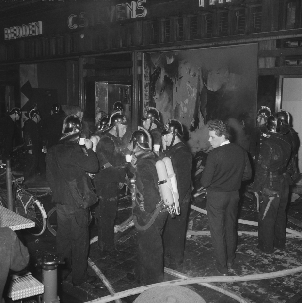 Schwarzes Bild von Feuerwehrleuten in Uniform mit Atemgeräten neben einem Gebäude, einem Fahrrad und einem Tisch mit Gegenständen.