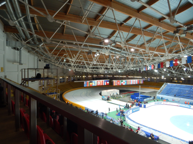Große Innen-Eislaufbahn voller Menschen, umgeben von Sitzplätzen, Geländern, Brettern, Leuchten und strukturellen Elementen, mit einer Wand im Hintergrund.