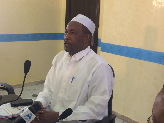 Ein Mann in einem weißen Kleid und Hut sitzt an einem Tisch mit Mikrofonen, spricht, während Papiere und Gegenstände auf dem Tisch liegen, mit einer anderen Person mit Brille rechts von ihm, in einem Raum mit einer Wand und einer Tür dahinter.