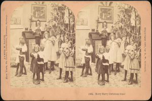 Gruppe von Kindern mit zwei Erwachsenen in der Mitte vor einem Weihnachtsbaum, umgeben von gerahmten Fotos an der Wand.