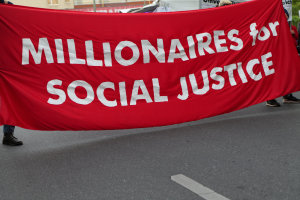 Eine Gruppe von Menschen marschiert mit einer roten Fahne, auf der "Millionaires for Social Justice" steht, eine Straße entlang mit Gebäuden, Strommasten, Bäumen und einem klaren blauen Himmel im Hintergrund.