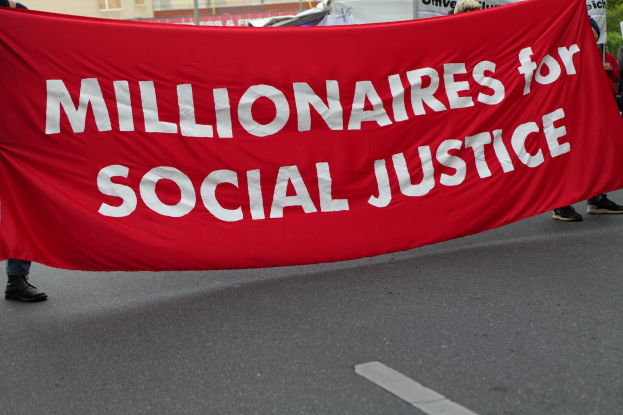 Eine Gruppe von Menschen marschiert mit einer roten Fahne, auf der "Millionaires for Social Justice" steht, eine Straße entlang mit Gebäuden, Strommasten, Bäumen und einem klaren blauen Himmel im Hintergrund.