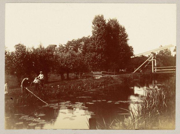 Ein altes Schwarz-Weiß-Foto von Menschen, die in einem Teich umgeben von Bäumen, Pflanzen und einem Holzzaun angeln, mit dem Himmel im Hintergrund.