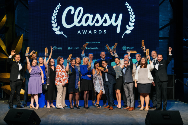 A group of people standing on a stage, smiling and holding awards, with speakers at the bottom and a screen in the background displaying "Classy Awards 2019".