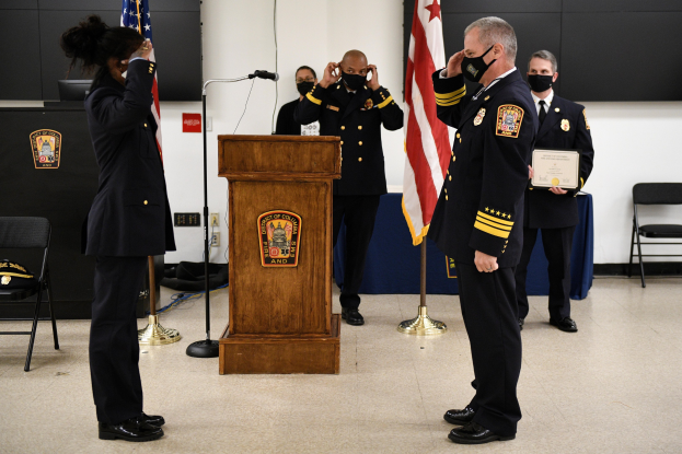 Gruppe uniformierter Personen in Masken vor einem Podium mit Mikrofon, umgeben von Fahnen und Möbeln, bei einer Vereidigung, bei der eine Person ein Zertifikat hält.