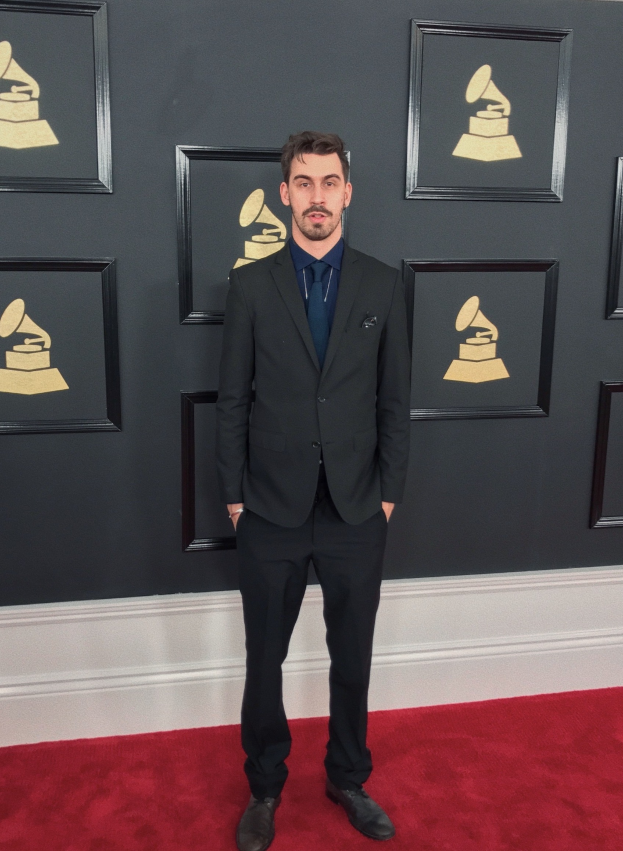 Ein Mann im Anzug steht auf dem roten Teppich bei den 55. Annual Grammy Awards, umgeben von einer Wand, die mit Bildern von der Preisverleihung geschmückt ist.