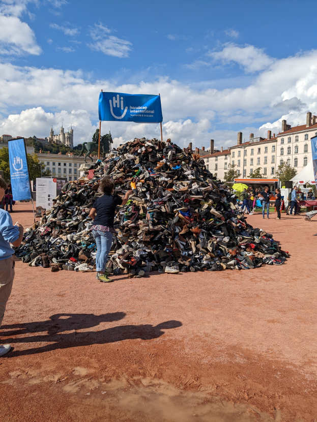 Eine Gruppe von Menschen steht um einen Haufen von Schuhen auf einem Schmutzfeld, mit Gebäuden, Bäumen und einem bewölkten Himmel im Hintergrund und Schildern mit Text, der auf ein internationales Schuhrecycling-Event hinweist.