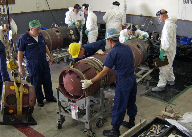 Gruppe von Männern in Helmen und Handschuhen, die eine große Bombe in einer Fabrik mit Maschinen und einer wandmontierten Textanzeige entschärfen.