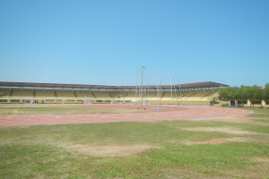 Großes Stadion mit einer zentralen Laufbahn, grünem Rasen, Bäumen auf der rechten Seite, Pfosten im Hintergrund und einem klaren blauen Himmel.