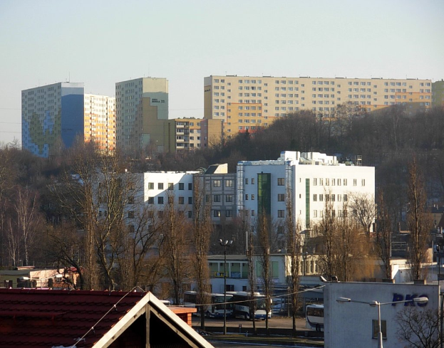Eine Stadtansicht von einem Hügel aus, mit Gebäuden, Bäumen, Straßenlaternen, Strommasten, Kabeln und einem klaren blauen Himmel.