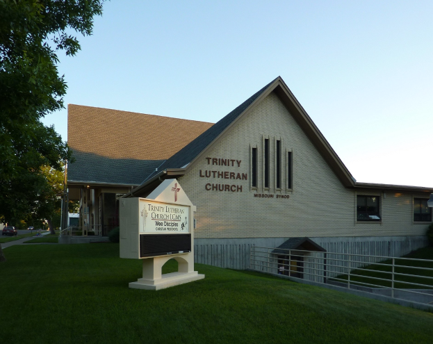 Außenansicht der Trinity Lutheran Church in St. Louis, Missouri, mit einem Gebäude mit Fenstern, einer Schautafel, einem Metallzaun, Gras, einem Straßenmast, Bäumen, Fahrzeugen auf der Straße und einem bewölkten Himmel.