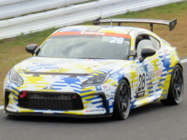 Ein weißer Toyota Supra GT3-Rennwagen mit Sponsor-Text und Nummern auf einer Strecke, umgeben von einer Begrenzung und Gras.