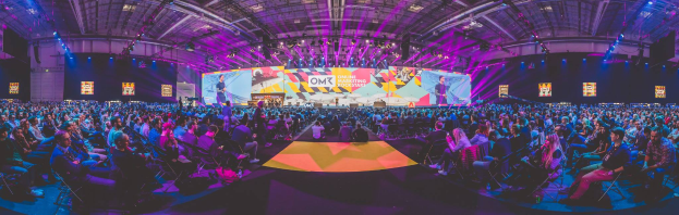 Eine große Menschenmenge sitzt auf Stühlen vor einer hell erleuchteten Bühne mit mehreren stehenden Individuen, wahrscheinlich bei einem Online-Marketing-Rockstars-Event, umgeben von Bildschirmen.