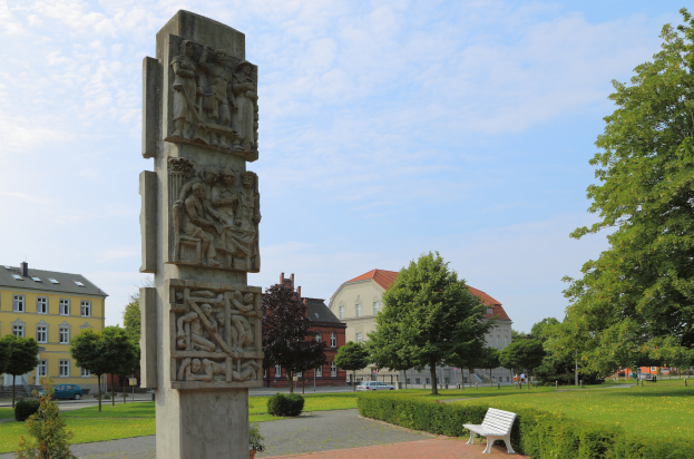Ein steinernes Denkmal in einem Park mit einer Bank davor, umgeben von grünem Gras, Pflanzen und Bäumen, mit Gebäuden, Fahrzeugen und einem bewölkten Himmel im Hintergrund.