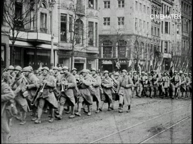 Schwarzes und weißes Foto von Soldaten, die während des Ersten Weltkriegs eine von Bäumen gesäumte Straße in Berlin entlangmarschieren, mit Mützen und Gewehren, sowie Gebäuden und einem Pfahl im Hintergrund.
