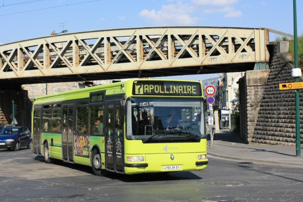 Grüner Bus fährt unter einer Brücke auf einer Stadtstraße mit Passagieren, umgeben von anderen Fahrzeugen, einem Pfahl mit Schildern, einer Wand, Gebäuden, Bäumen und einem bewölkten Himmel.