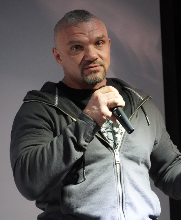 Ein Mann in einer schwarzen Kapuzenjacke und einem schwarzen Anzug mit einem weißen Hemd, der selbstbewusst vor einer Wand steht und ein Mikrofon hält, sieht aus wie ein Komiker mit zurückgekämmten Haaren.