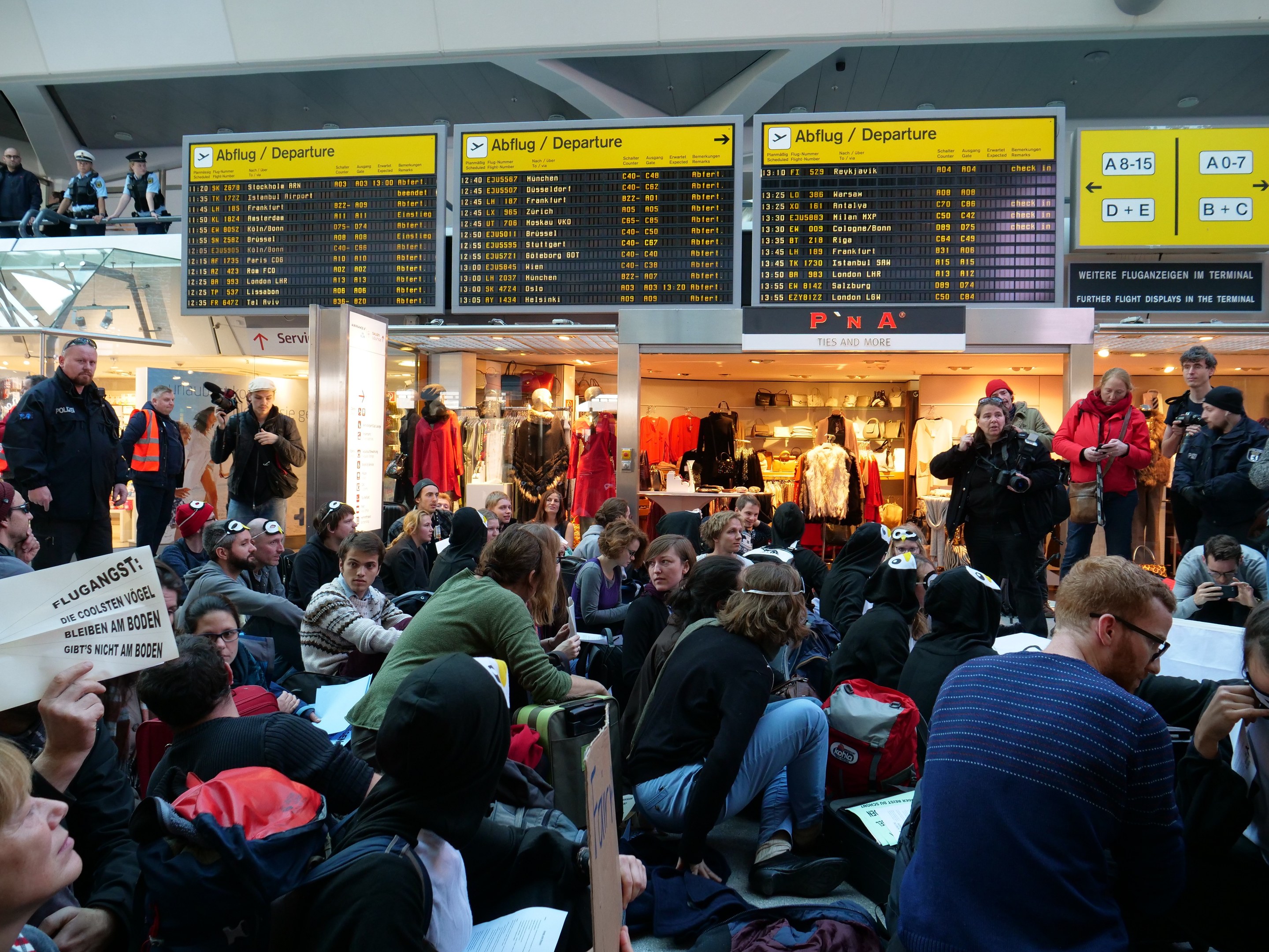 Eine große Gruppe von Menschen an einem Flughafen, einige sitzen mit Taschen und Papieren, andere stehen, mit Texttafeln, Schaufensterpuppen in Kleidern und Deckenbeleuchtung im Hintergrund, was auf eine Demonstration hinweist.