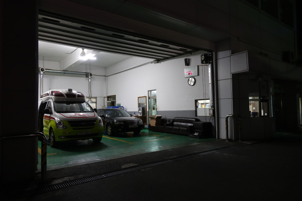 Ambulanz in einem Gebäude bei Nacht mit sichtbaren Fahrzeugen, Sofas, einer Wanduhr, einer Texttafel, einer Tür, Rohren, Deckenlampen und Metallstangen.