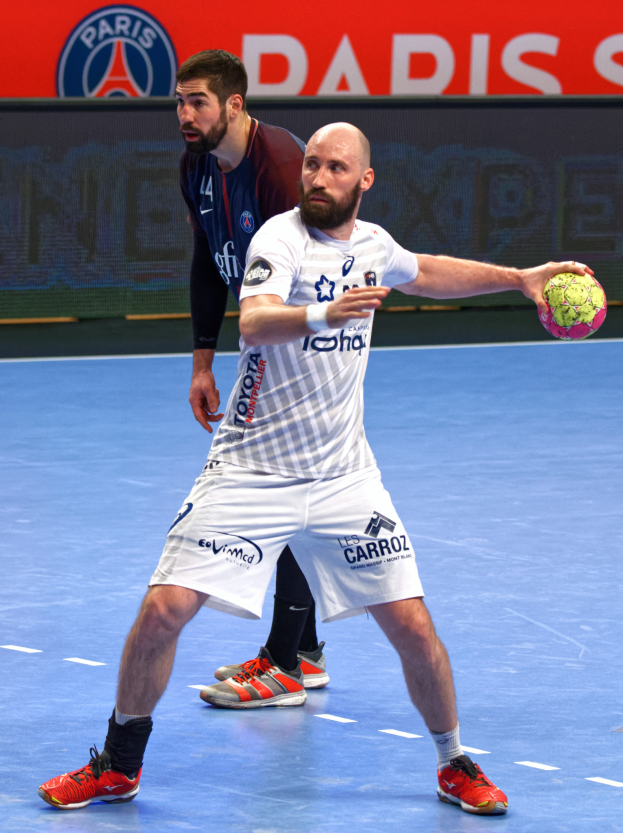 Zwei Männer spielen Handball auf einem Platz, einer hält einen Ball, mit einer Tafel im Hintergrund, die 'Paris Saint-Germain vs Paris Saint Germain' anzeigt.