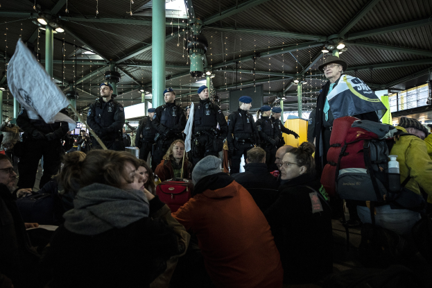 Eine Gruppe von Menschen steht vor einer größeren Menge, die scheinbar ein Bahnhof ist, wobei einige Personen Mützen tragen, Taschen tragen und Protestschilder halten.