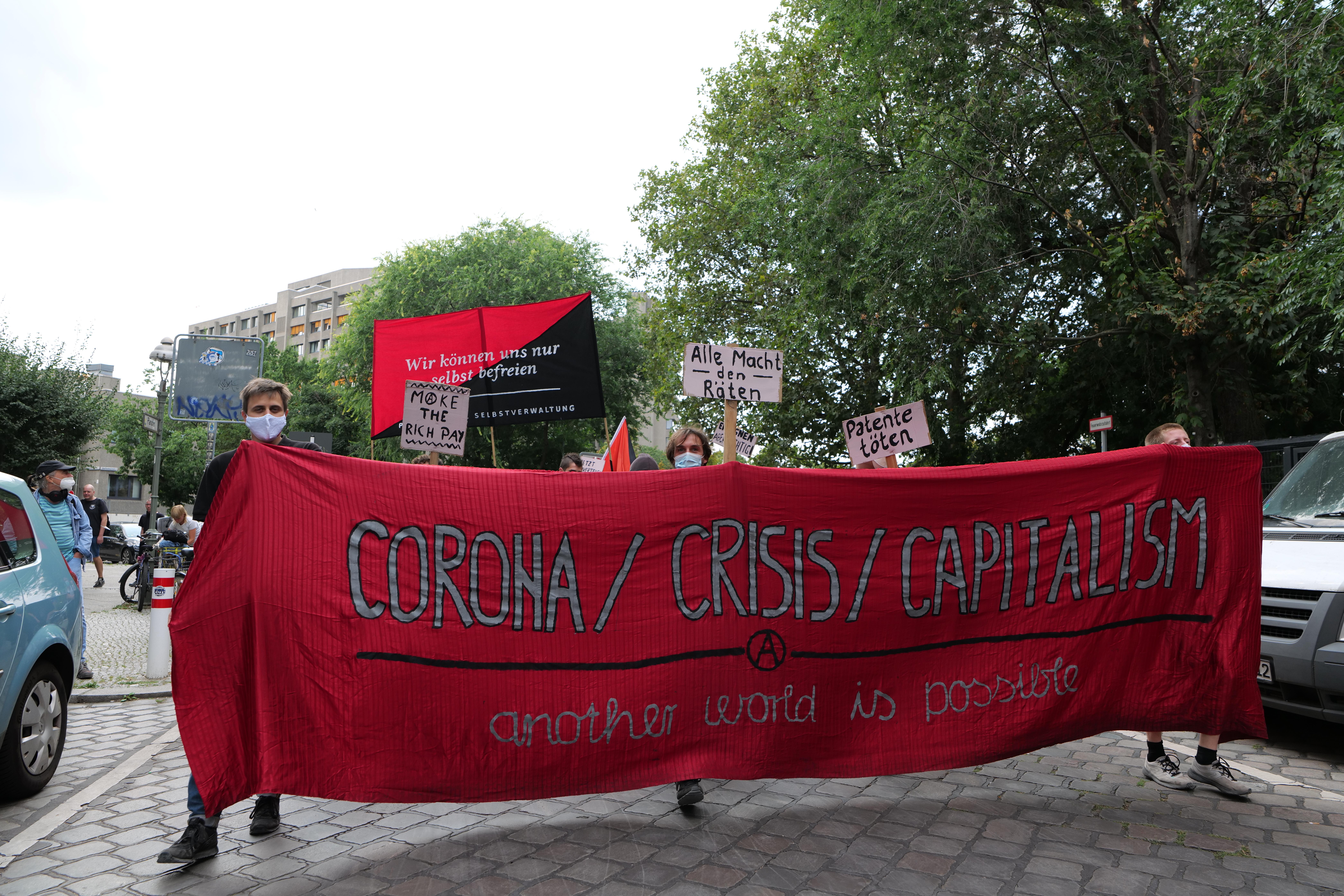 Eine Gruppe von Menschen marschiert mit einer roten Fahne, auf der "Corona-Krise/Kapitalismus" steht, eine Straße entlang. Im Hintergrund sind geparkte Fahrzeuge, Gebäude und Bäume zu sehen.