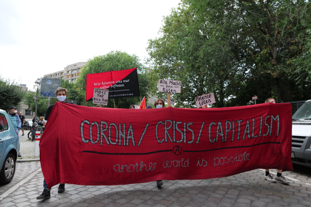 Eine Gruppe von Menschen marschiert mit einer roten Fahne, auf der "Corona-Krise/Kapitalismus" steht, eine Straße entlang. Im Hintergrund sind geparkte Fahrzeuge, Gebäude und Bäume zu sehen.
