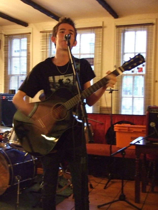 Ein Mann in einem schwarzen T-Shirt und Hose steht mit einer Gitarre und einem Mikrofon auf einem Ständer, mit einer blauen Trommel in der Ecke, einer Couch mit Gegenständen darauf und durchsichtigen Vorhängen im Hintergrund.