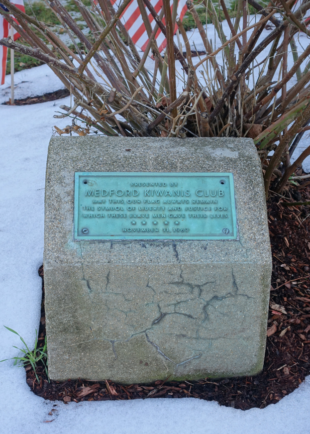 Ein Gedenkstein mit einer Plakette, die 'Medford Kiwanis Club' trägt, im Schnee umgeben von Pflanzen und einer Flagge im Hintergrund.