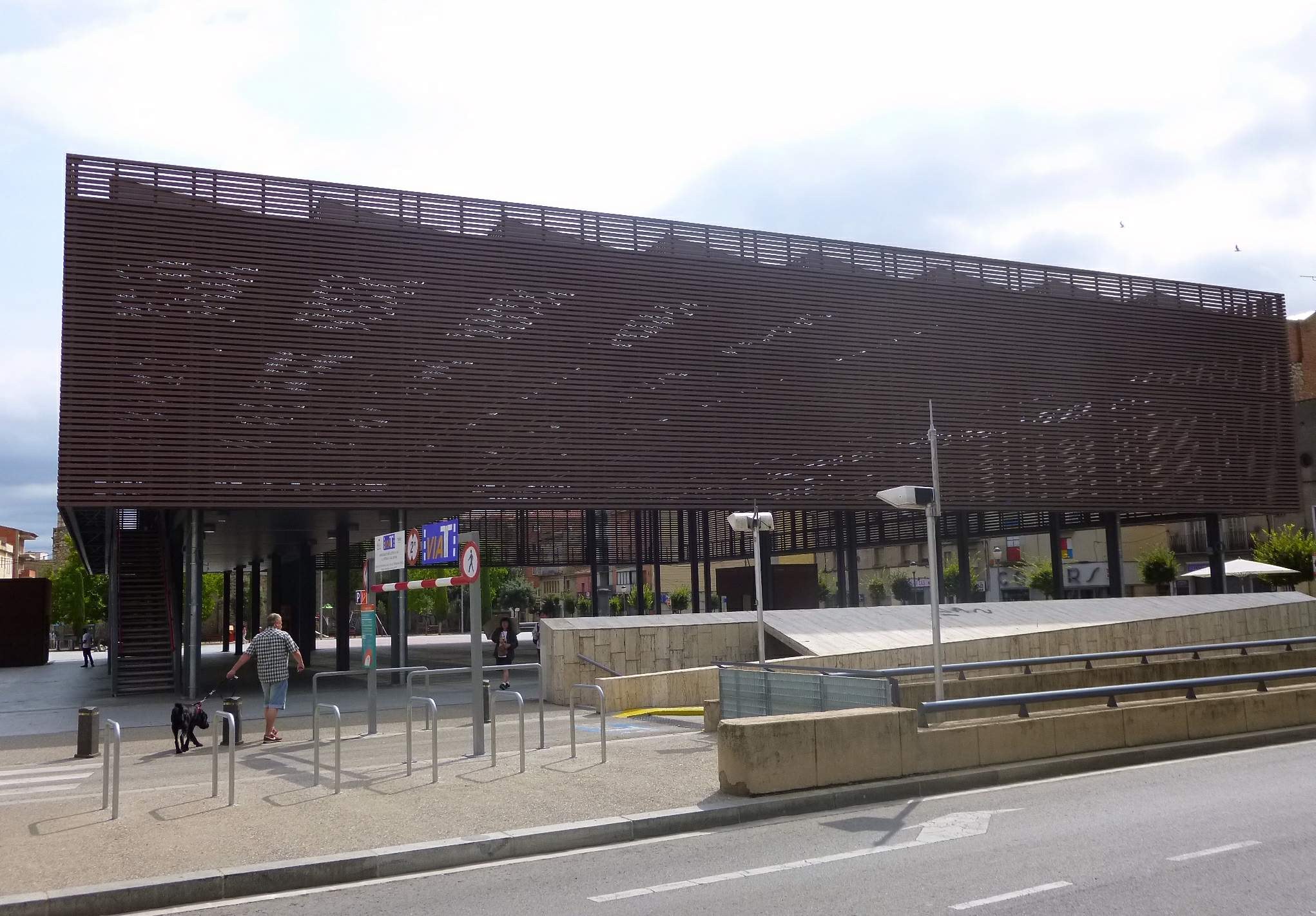 Großes Gebäude mit zahlreichen Fenstern, das den neuen Eingang zur Stadtmitte bildet, umgeben von Straßeninfrastruktur, Fahrzeugen, Fußgängern, Bäumen und einem bewölkten Himmel.
