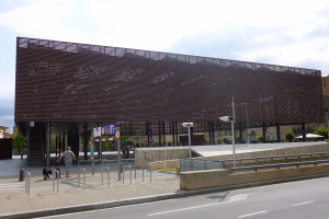 Großes Gebäude mit zahlreichen Fenstern, das den neuen Eingang zur Stadtmitte bildet, umgeben von Straßeninfrastruktur, Fahrzeugen, Fußgängern, Bäumen und einem bewölkten Himmel.