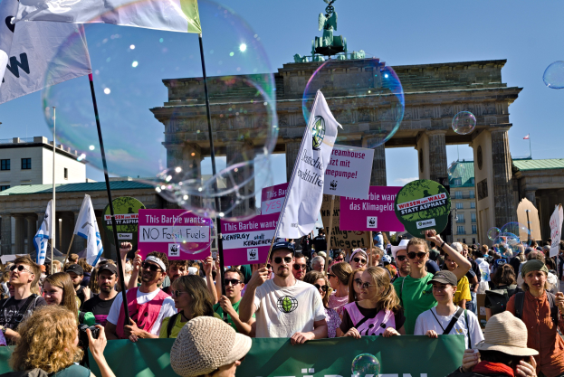 Menschenmenge vor dem Brandenburger Tor in Berlin, viele mit Mützen und Schutzbrillen, einige mit Fahnen und Schildern mit Text, im Hintergrund Gebäude, ein von einer Statue gekrönter Bogen und ein mit Seifenblasen gefüllter Himmel.