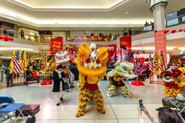 Ein lebendiges chinesisches Neujahrsfest in einem Einkaufszentrum mit tanzenden Menschen in Kostümen, anderen auf Stühlen sitzend mit Mobiltelefonen und verschiedenen Dekorationen, Fahnen und Bannern im Hintergrund.