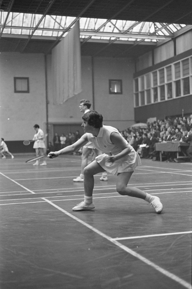 Schwarzes und weißes Foto von zwei Frauen, die Badminton auf einem Platz spielen, jede hält einen Schläger, mit Zuschauern rechts und einer Wand mit Fenstern im Hintergrund.