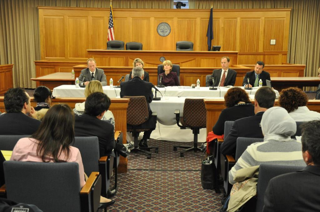 Ein Versammlungsraum mit einer Gruppe von Menschen, die auf Stühlen sitzen, zwei ausländische Flaggen, ein oranger Schreibtisch mit Wasserflaschen und einem Mikrofon.