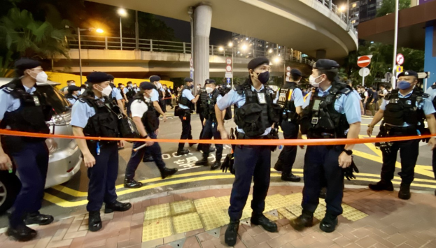Eine Gruppe von Polizisten steht vor einer Menge uniformierter Personen mit Masken, einem Band im Vordergrund und Fahrzeugen links, mit Gebäuden, Bäumen, Laternenmasten, Schildern und einer Brücke mit Geländern im Hintergrund, wahrscheinlich zeigt es die Nachwehen einer Hongkonger Protestaktion.