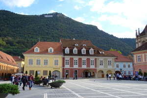 Gruppe von Menschen, die durch einen Stadtplatz mit Gebäuden, Fenstern, Laternenmasten und Topfpflanzen sowie einem Hügel mit Bäumen und einem bewölkten Himmel gehen, mit Text auf einem Gebäude im Hintergrund.