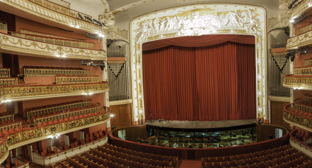 Großer Zuschauerraum mit roter Gardine, Sitzreihen, Geländern, Beleuchtungskörpern und Wandskulpturen, mit Bühnenobjekten, die eine Konzerthalle suggerieren.