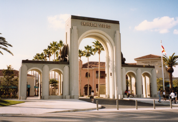 Eingang von Universal Studios Hollywood mit einem Bogen, einer Statue, Menschen, Bäumen, Gebäuden, einem Fahnenmast und einem bewölkten Himmel.