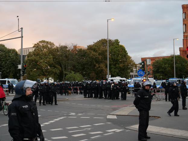 Eine Gruppe von Polizisten in Helmen steht in der Mitte einer Straße umgeben von Fahrzeugen, Verkehrsampeln, Laternen, Bäumen, Gebäuden mit Fenstern und einem bewölkten Himmel.