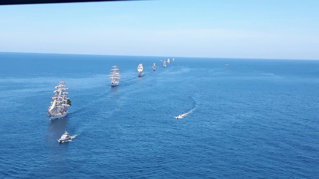 Eine große Gruppe von Segelschiffen auf dem Meer während eines Regattas, mit einem klaren blauen Himmel und hellen Sonnenstrahlen, die sich auf dem Wasser spiegeln.