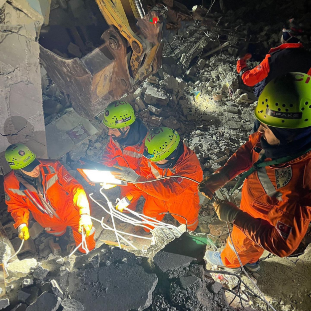 Gruppe von Menschen in orangefarbenen Uniformen und grünen Helmen bei der Arbeit an Schutt mit einem Kran im Hintergrund, hölende Rettungsgeräte.